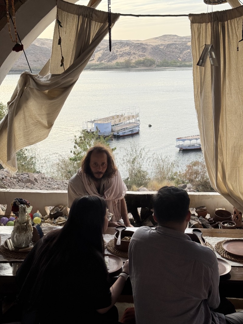 George at the low table with guests, the Nile and the boats behind him