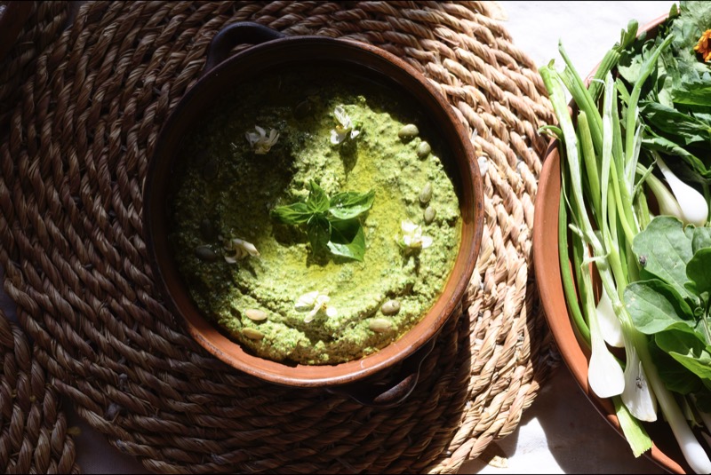 A bowl of green herb dip with spring onions and fresh leaves, on a woven mat